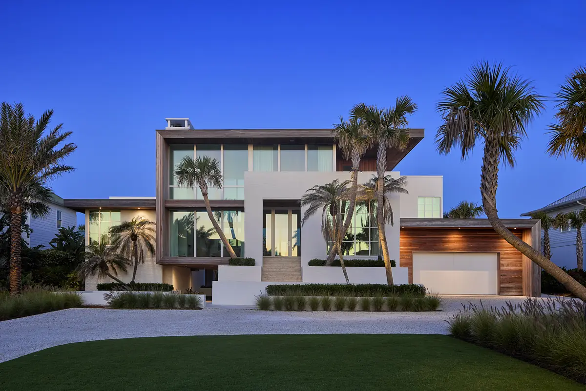 Modern house with white stucco, wood elements, large windows, driveway, palm trees, and green landscaping