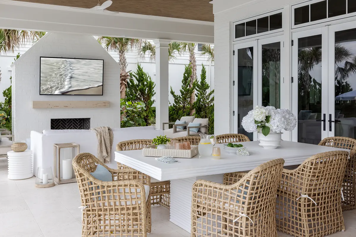 Outdoor deck with white table, wicker chairs, mounted TV, stone fireplace, and greenery.