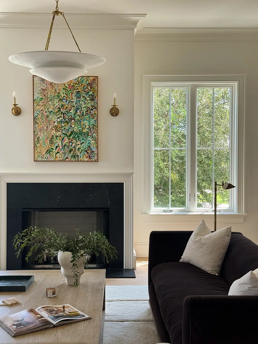 Living room with dark sofa, coffee table, side table, artwork, marble fireplace, and large windows with greenery view.