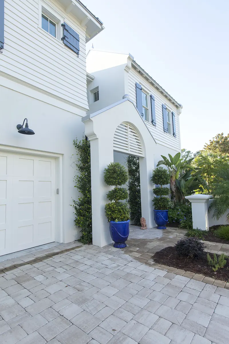 Exterior front view of a white house with blue shutters, archway, stone pathway, potted plants, and garage door
