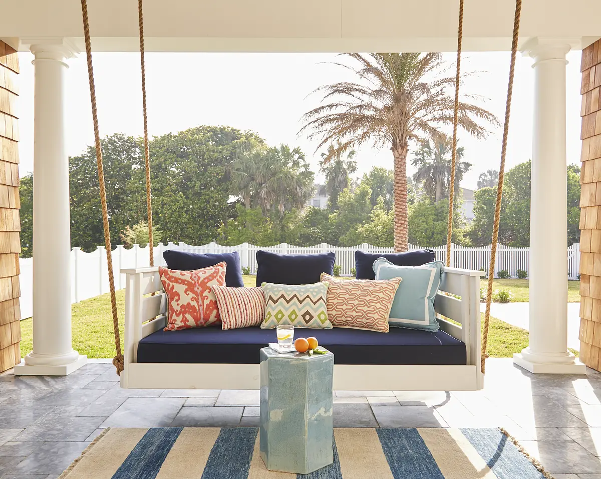 Patio with swing couch, blue cushions, colorful pillows, blue side table, and striped rug surrounded by greenery