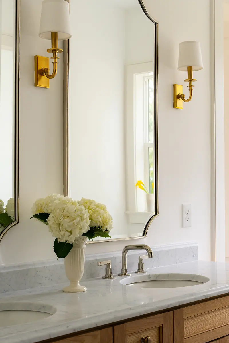 Bathroom with double sink vanity, marble countertop, large mirror, white flowers, and gold wall sconces.