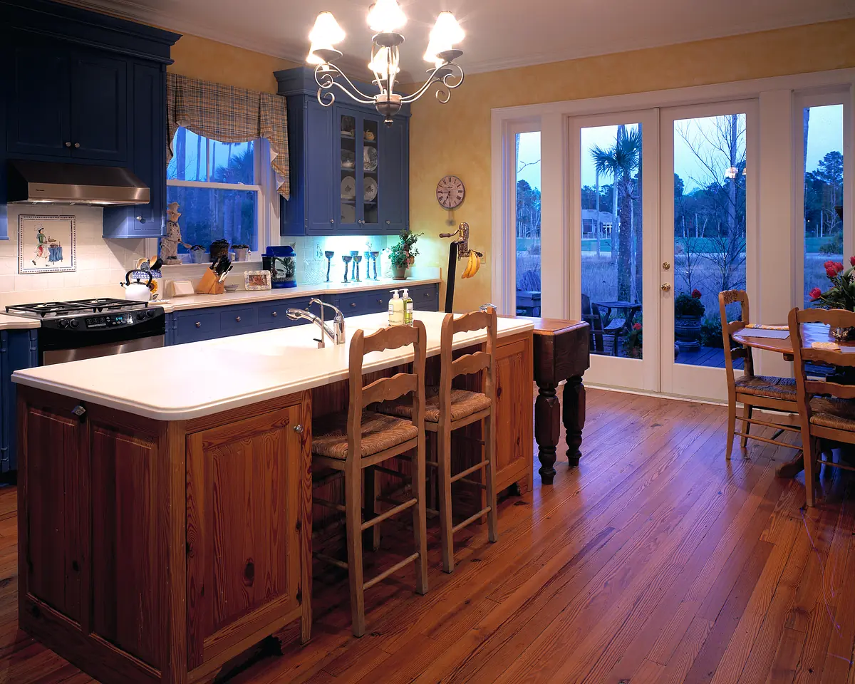 Kitchen with blue cabinets, white countertop, bar stools, wooden dining table, and large glass doors.