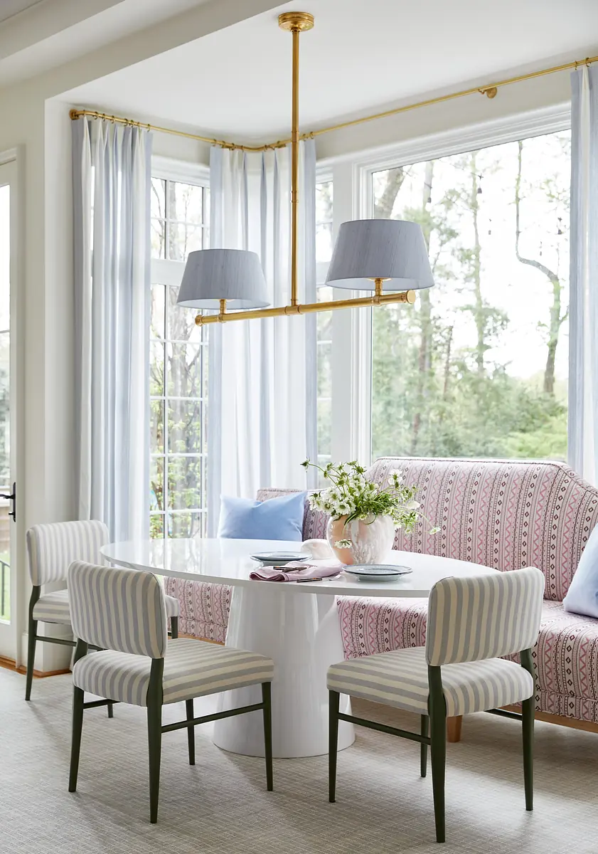 Dining room with white circular table, striped chairs, banquette, and large windows.