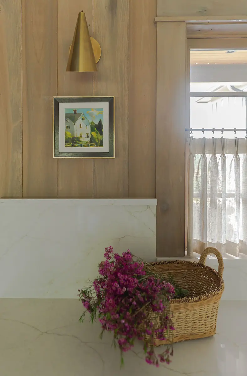 Kitchen area with marble countertop, wooden paneling, brass sconce, framed painting, and woven basket with flowers.