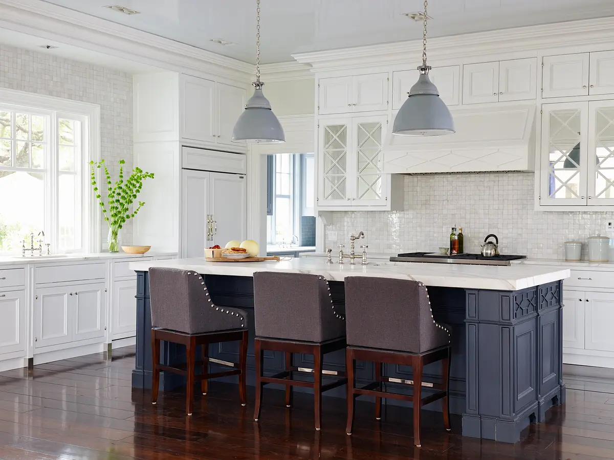 Kitchen with marble island, dark blue base, white cabinetry, three bar stools, and a large window with a plant.