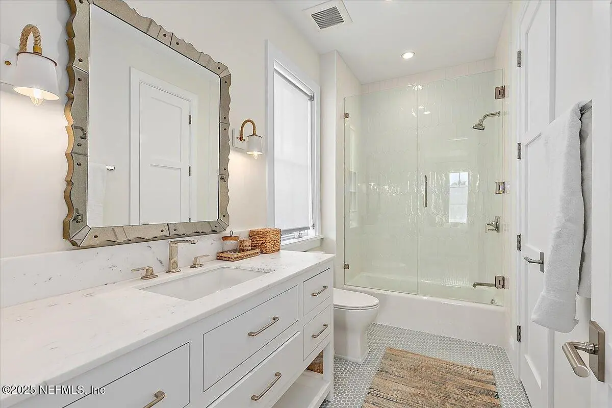 Bathroom with double vanity, bathtub with glass enclosure, toilet, and natural light from window.
