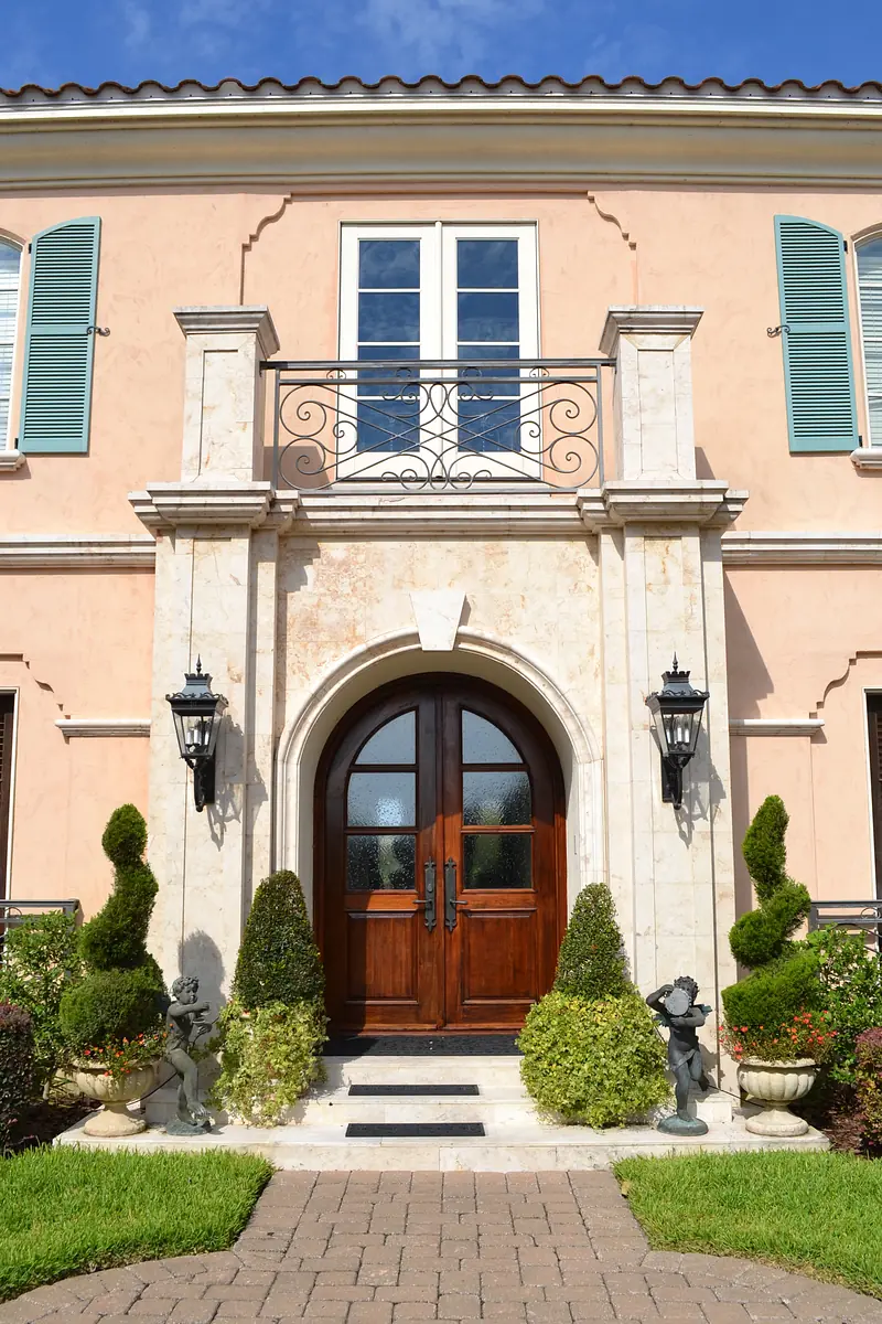 Front exterior with wooden door, lanterns, topiary bushes, planters, pink walls, and balcony with iron railing.