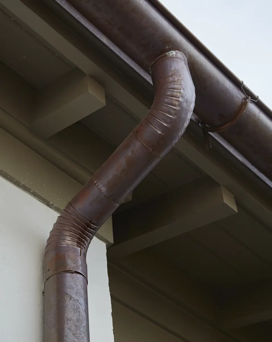 Close-up of brown metal downspout with corrugated texture beneath roof eave and light-colored wall.
