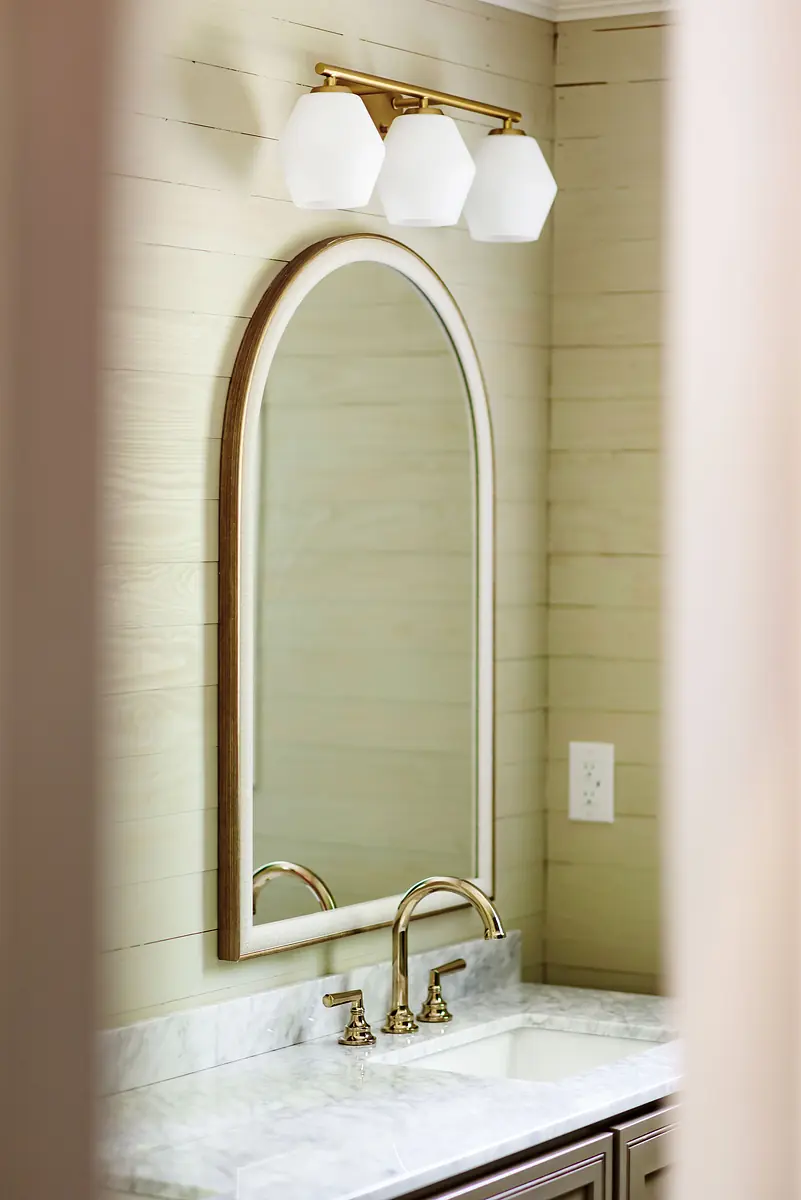 Powder room with marble countertop, arched mirror, gold faucet, and wooden panel walls.