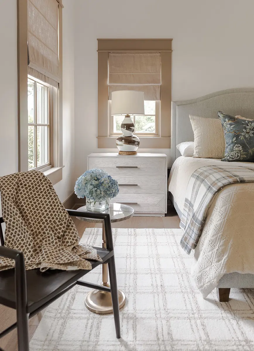 Bedroom with light gray bed, beige bedding, nightstand, chair, blue hydrangeas, and windows with beige shades