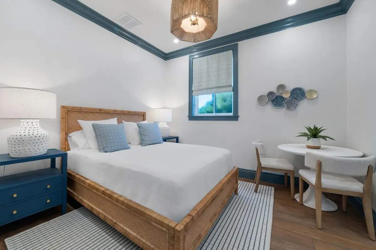 Bedroom with wooden bed, white quilt, blue pillows, round table, two chairs, and blue trim.