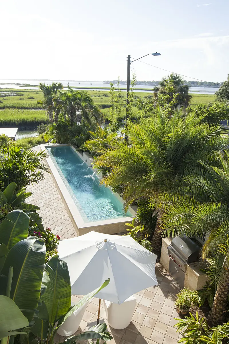 Patio with swimming pool, umbrella, tropical plants, and grill surrounded by tiled flooring.