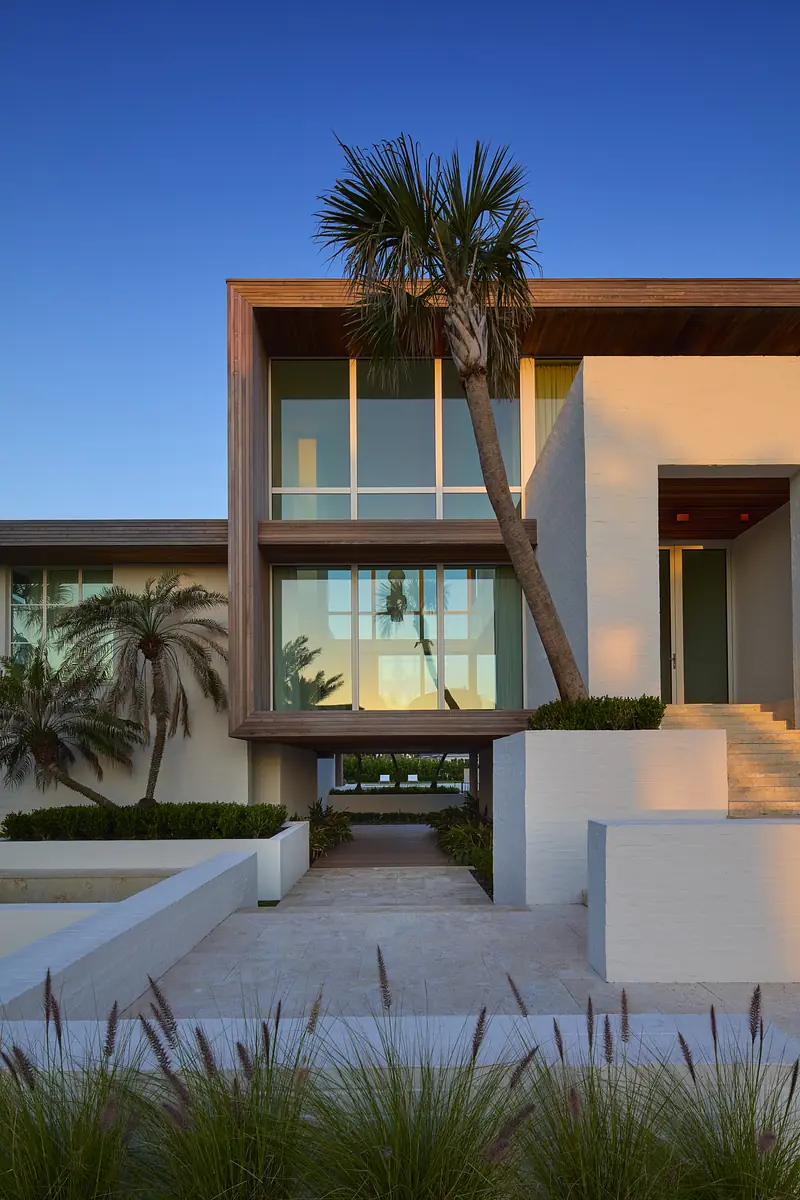 Modern house façade with large windows, wooden overhang, and stone steps surrounded by greenery.