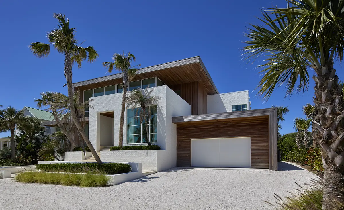 Modern house exterior with large windows, white walls, wooden accent, and palm trees.