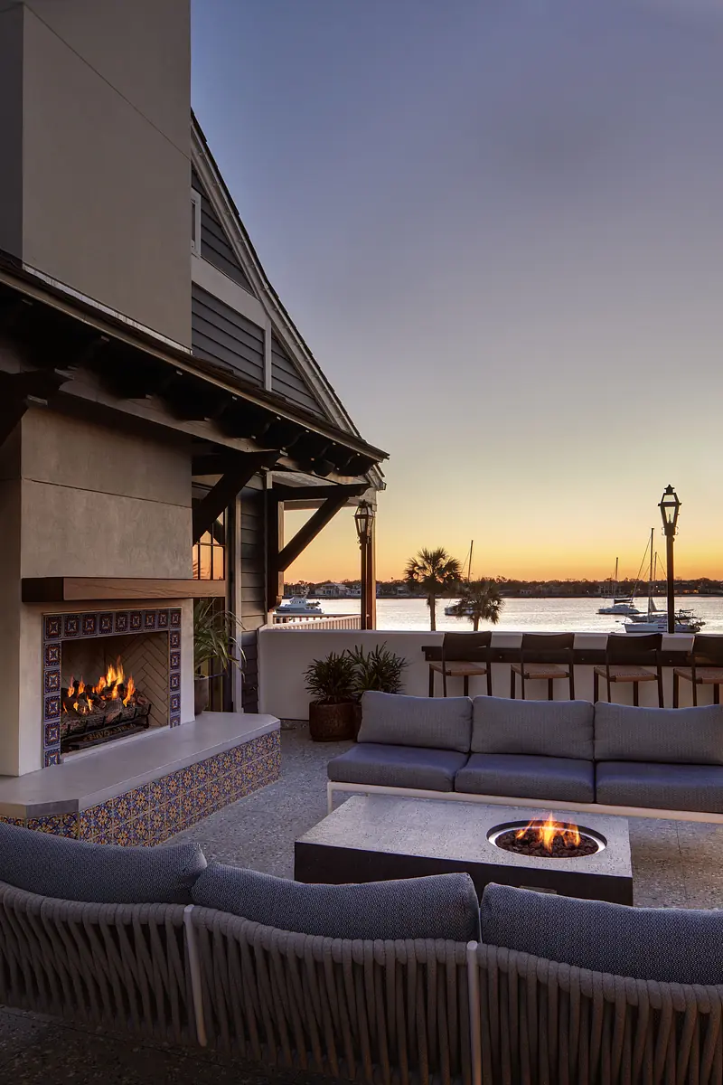 Deck with sectional sofa, fire pit table, tiled fireplace, and views of watercraft at sunset.