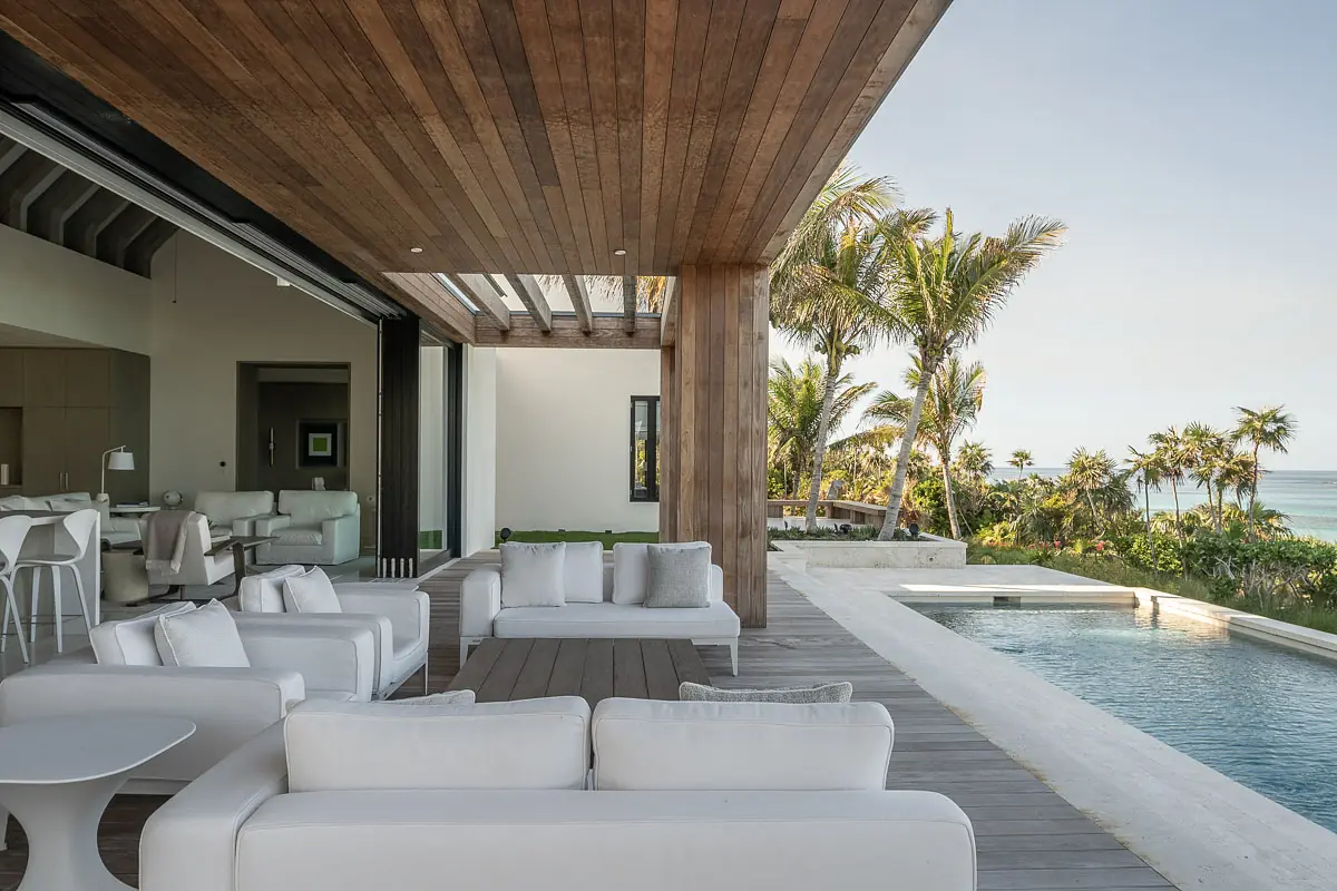 Patio area with white outdoor sofas, side table, swimming pool, and tropical landscaping under wooden beams