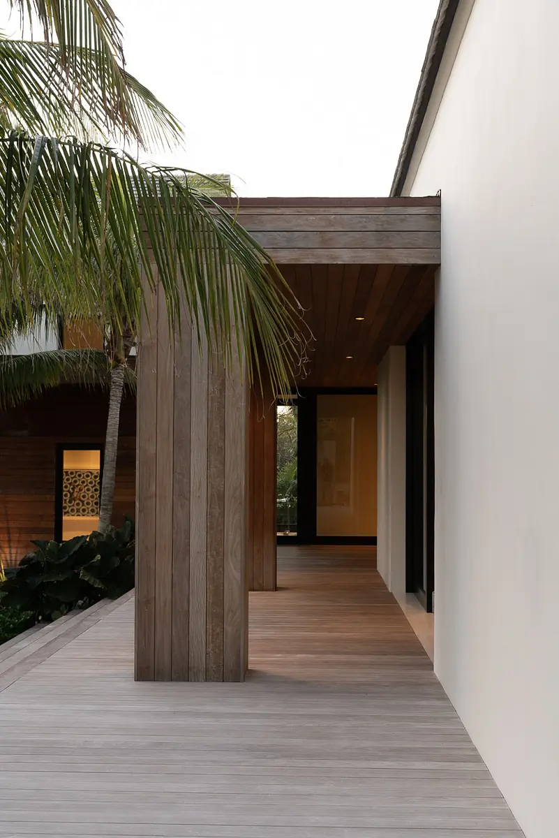 Exterior front view with wooden walkway, white walls, and tropical plants leading to a glass door.