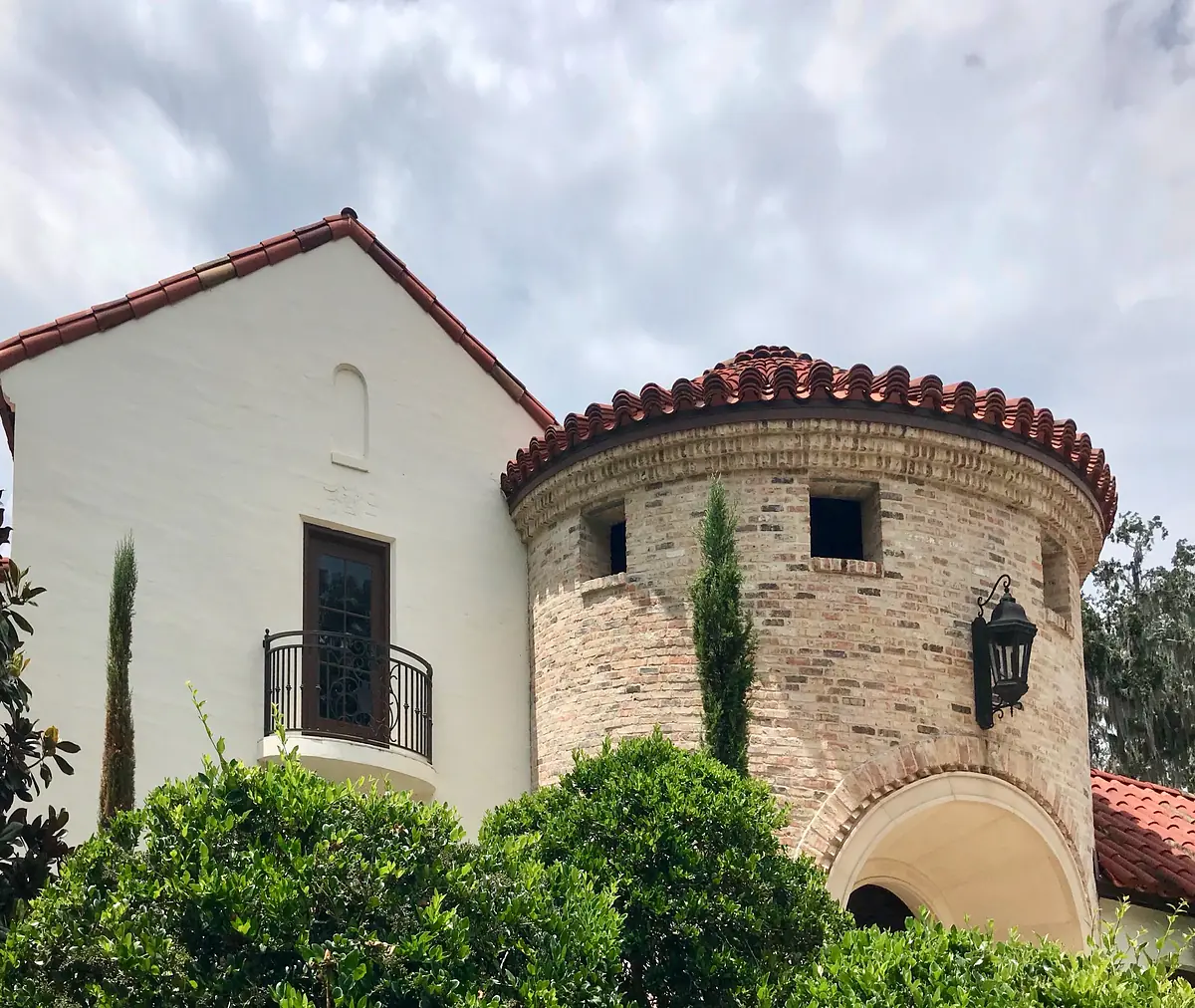 Exterior front view of a house with a round brick tower, red tile roof, balcony, and surrounding greenery.