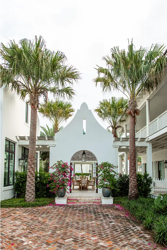 Exterior entrance with white archway, palm trees, flowering plants, and a brick path leading to a seating area.