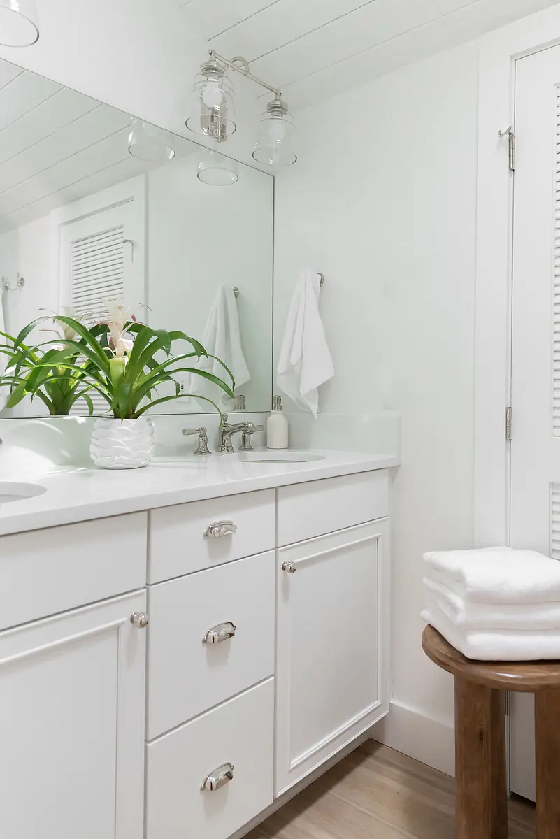 Bathroom with white vanity, large mirror, orchid plant, towel bars, wooden stool, and light wood flooring.