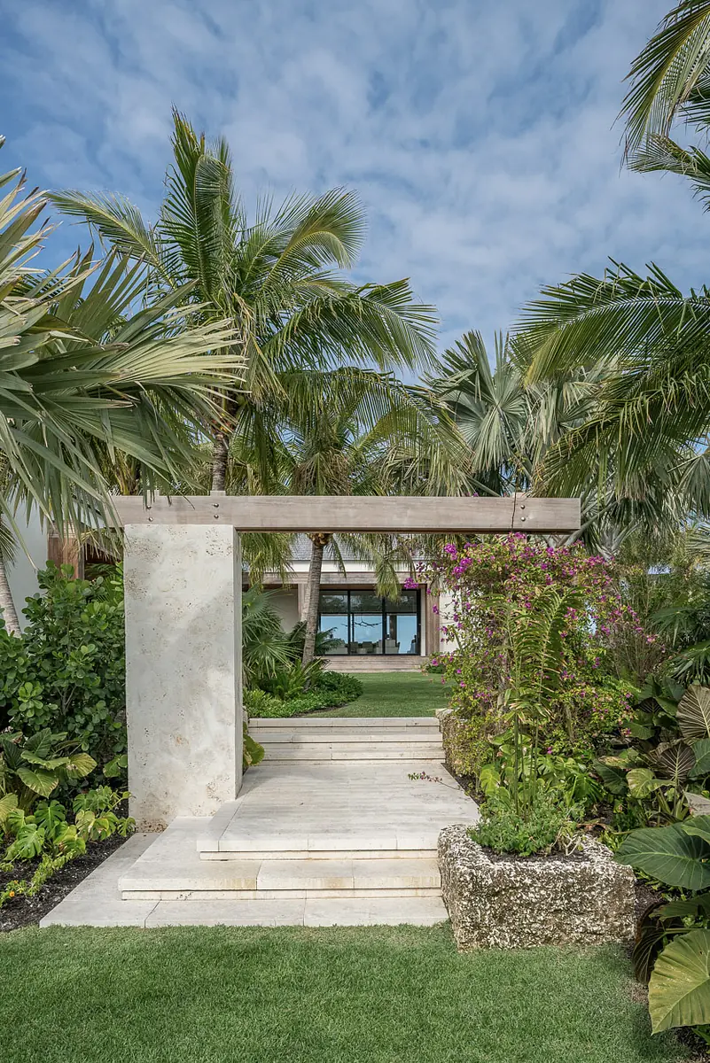 Exterior front view with stone pathway, palm trees, flowering plants, and modern building in background