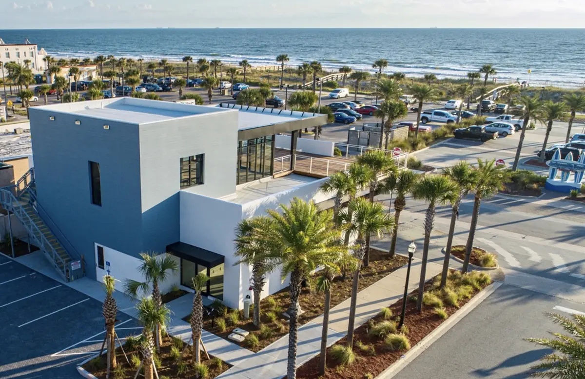 Exterior view of modern building with palm trees, parking lot, and ocean in background.