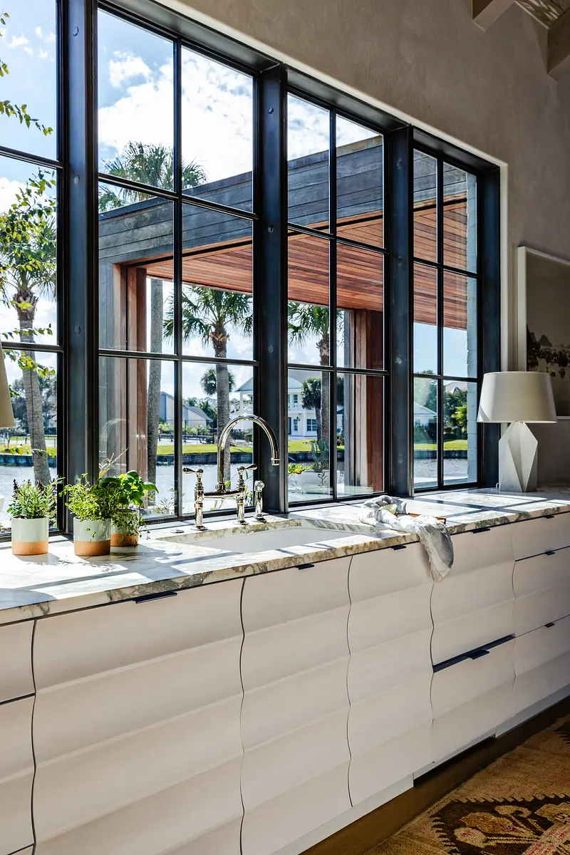 Kitchen with textured white cabinets, marble countertop, large windows, and potted herbs.