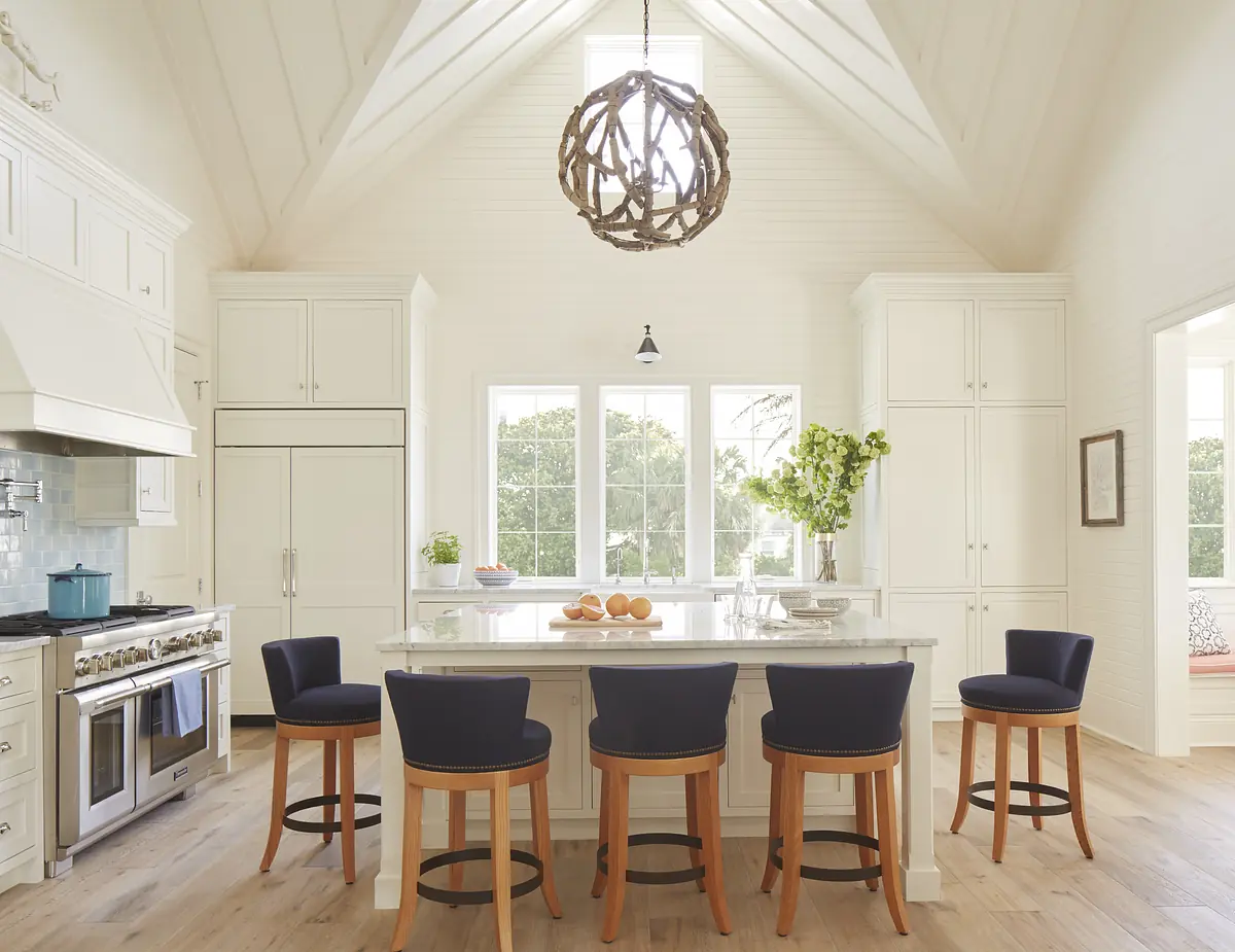 Kitchen with large island, stools, white cabinetry, marble countertops, stainless steel oven, and decorative light fixture.