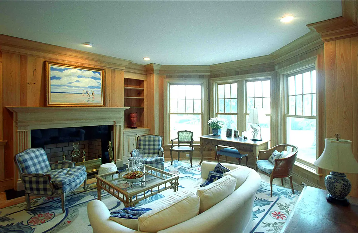 Living room with white sofa, glass coffee table, area rug, armchairs, bookcase, and bay windows with a fireplace