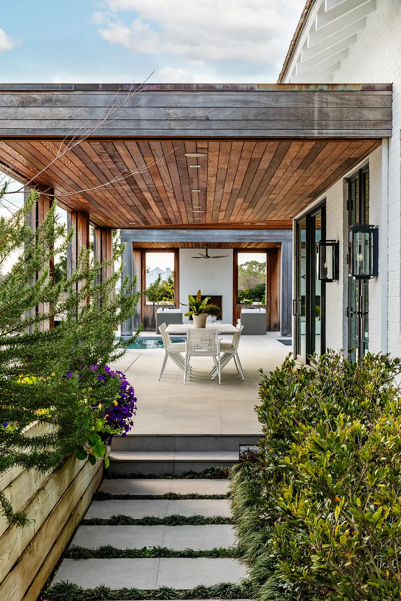 Patio with wooden ceiling, dining table, chairs, potted plants, and visible swimming pool in the background