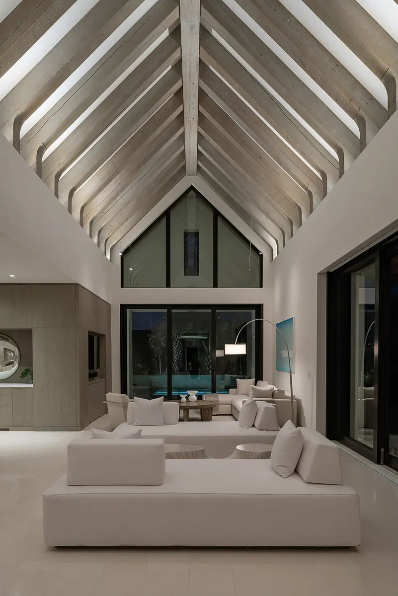 Living room with white sectional sofa, round coffee table, large windows, and exposed wooden beams.