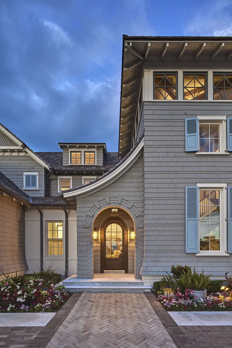 Exterior front with a large arched entry, gray shingle siding, blue shutters, and landscaped flower beds.