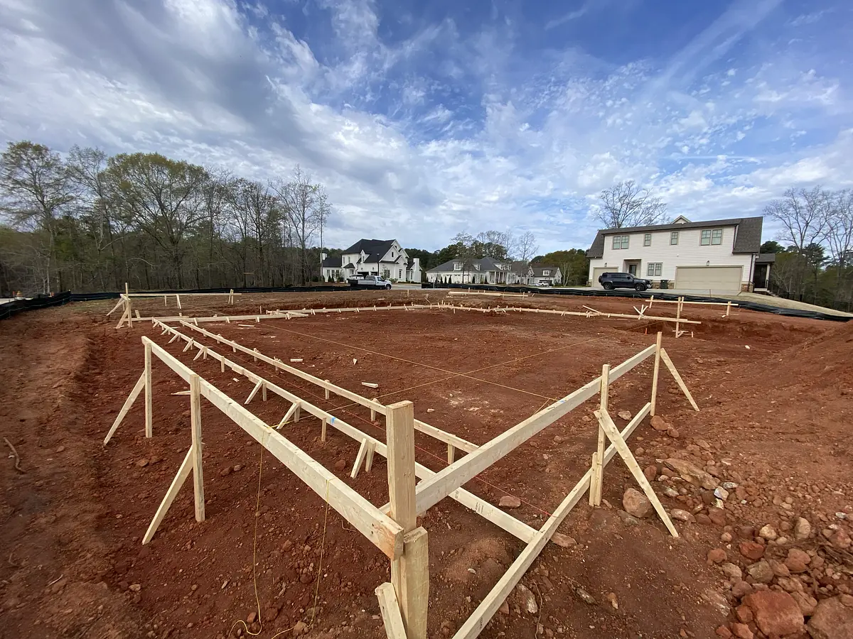 Construction site with wooden framing, orange-red dirt, and houses in the background.