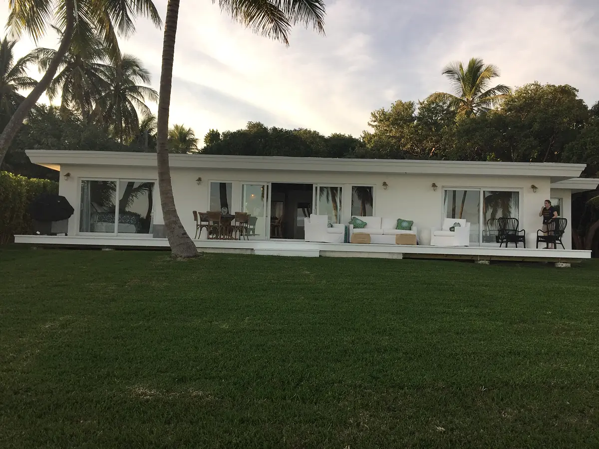 Exterior rear view of a house with large windows, patio seating, and palm trees.