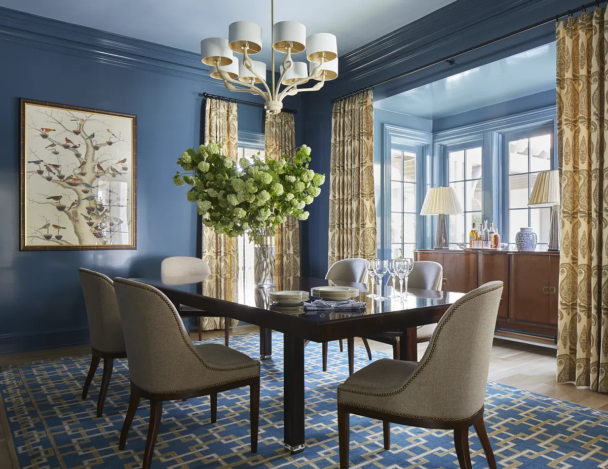 Dining room with dark wood table, upholstered chairs, blue walls, floral arrangement, and sideboard with lamps.
