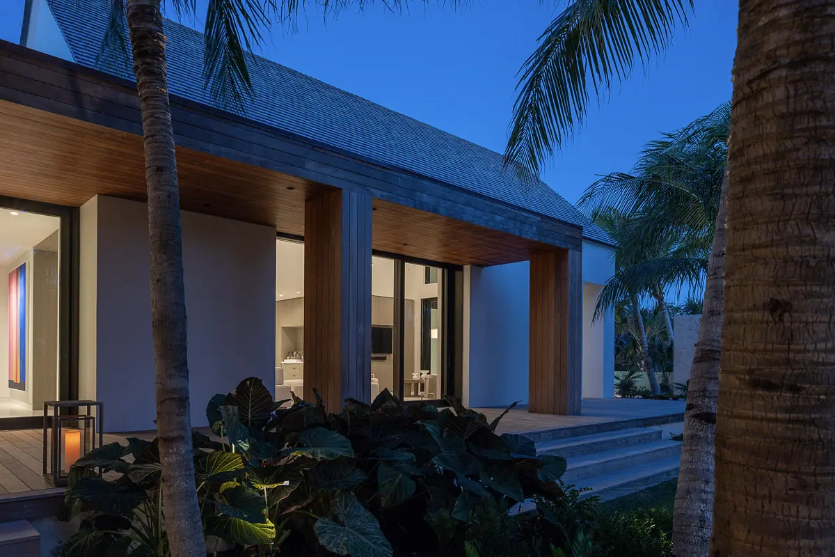 Exterior front view with sloped roof, wooden columns, glass doors, palm trees, and stone pathway.
