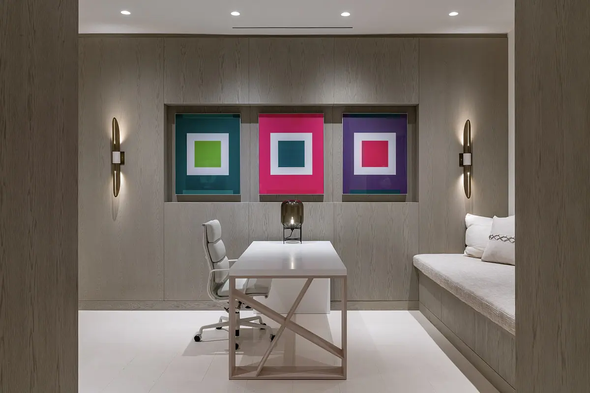 Office with white desk, office chair, built-in bench, colorful artworks, wood-textured walls, and wall sconces