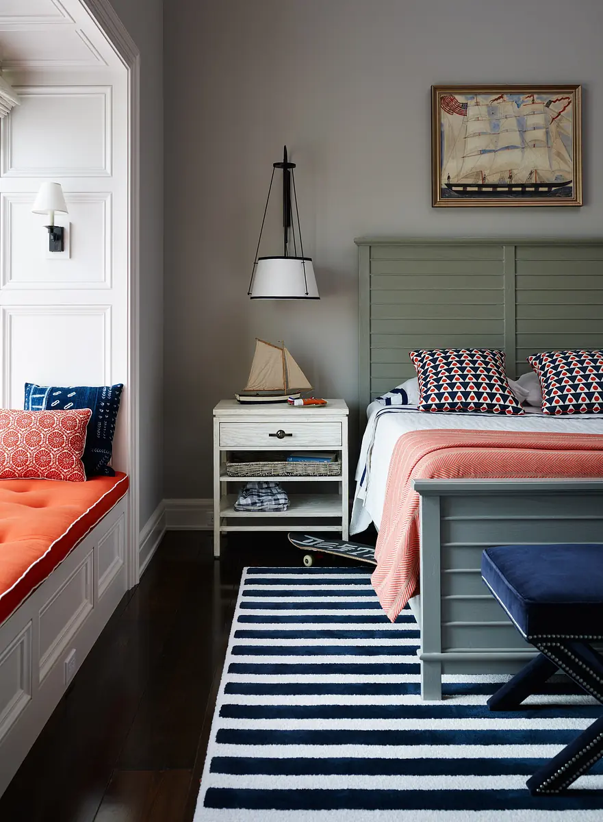 Bedroom with gray headboard bed, orange blanket, nightstand, striped rug, and window seat with cushions.