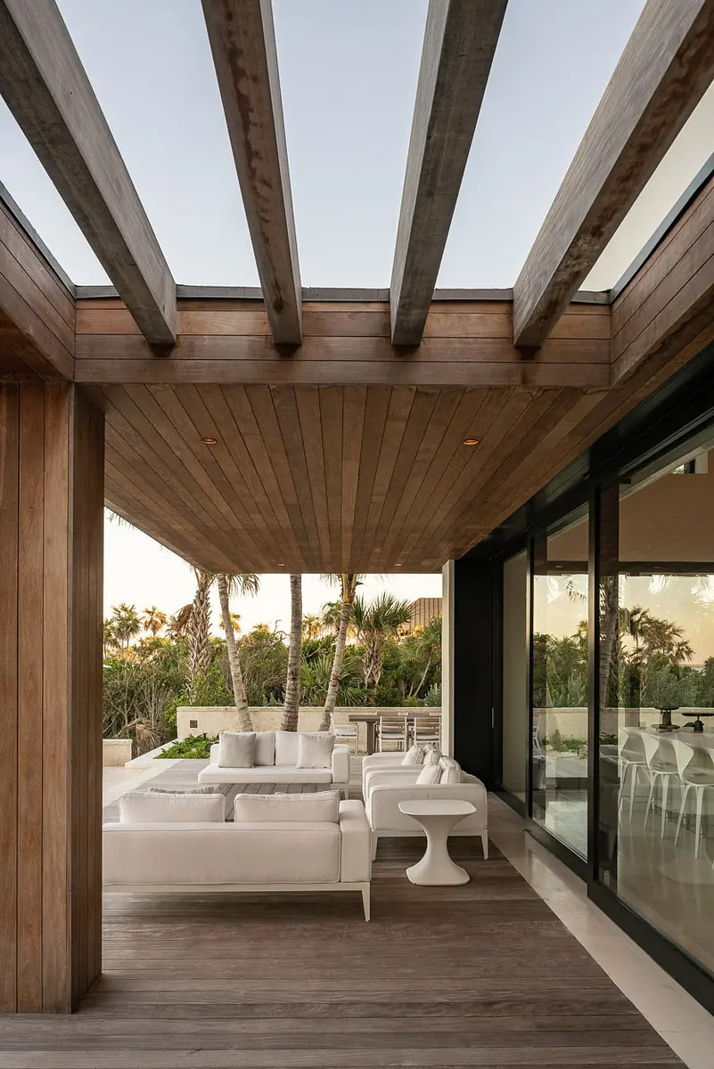 Patio with white seating, round side table, wooden beams, and large glass sliding doors with greenery in the background