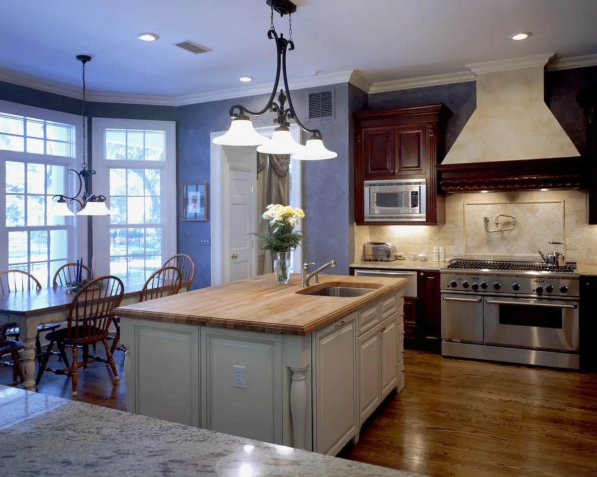Kitchen with wooden island, stainless steel appliances, wooden cabinets, and dining table beside large windows.