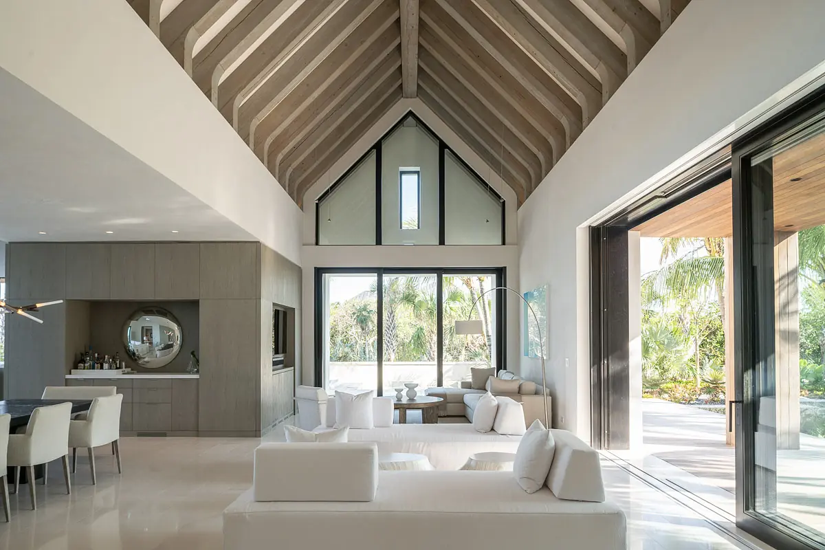 Living room with vaulted ceiling, large windows, neutral sofas, and circular coffee table leading to an outdoor area.