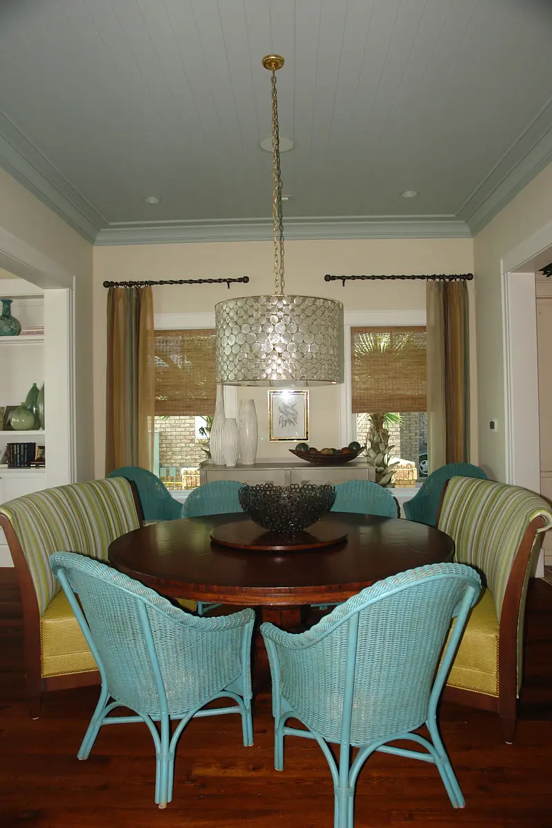 Dining room with round table, rattan chairs, decorative bowl, and large windows with woven shades.
