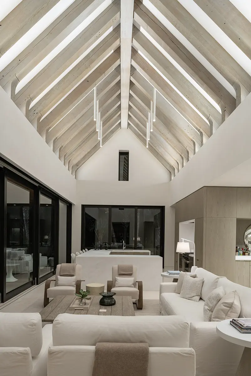 Living room with white sofas, wood coffee table, large windows, sloped ceiling, and bar area in background.