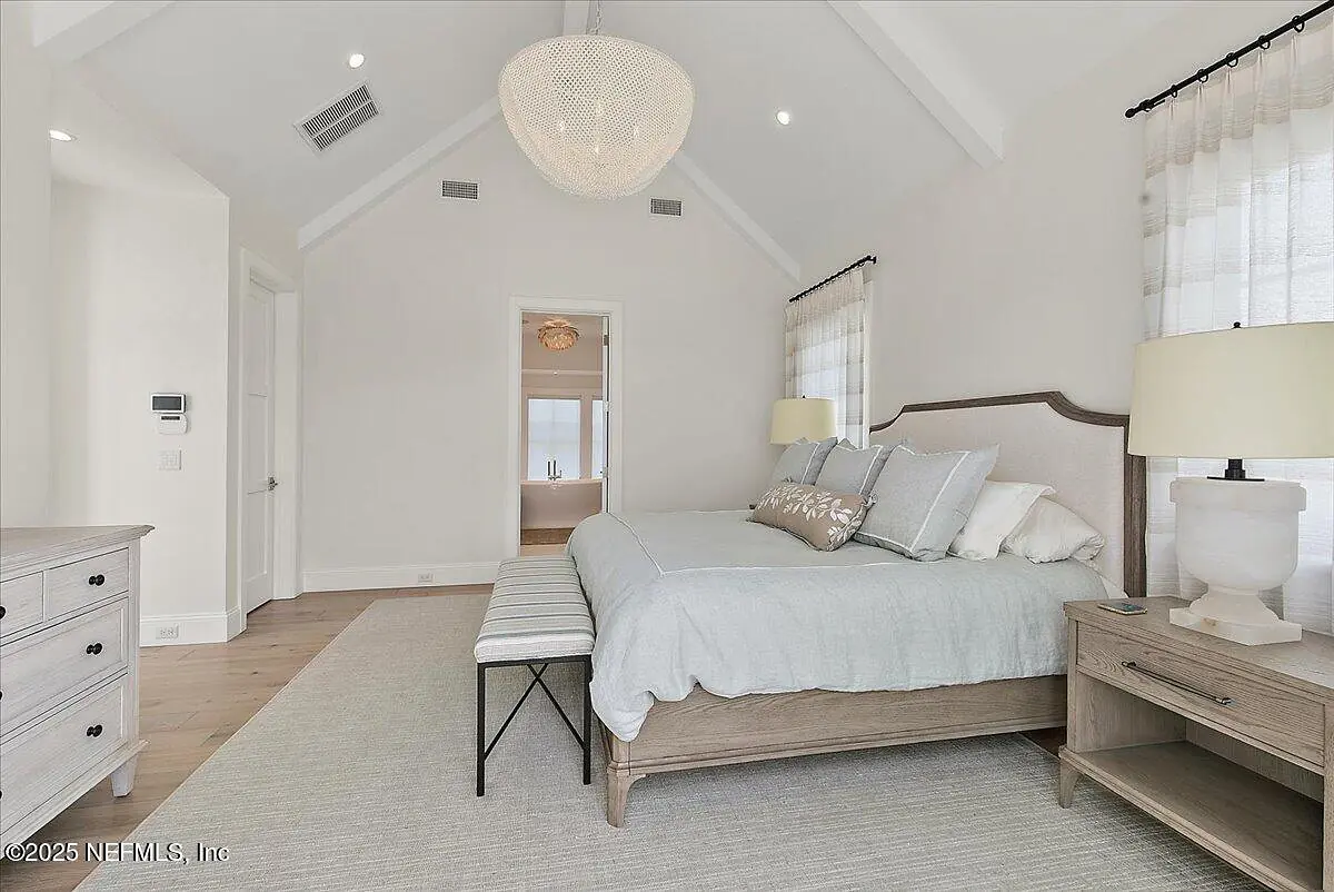 Bedroom with gray bed, wooden nightstand, chest of drawers, bench, and large windows with sheer curtains