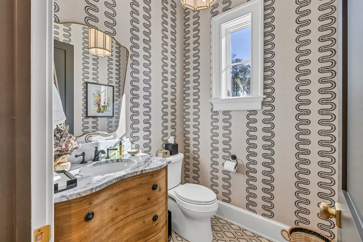 Powder room with wooden vanity, sink, toilet, round mirror, patterned wallpaper, and a small window.