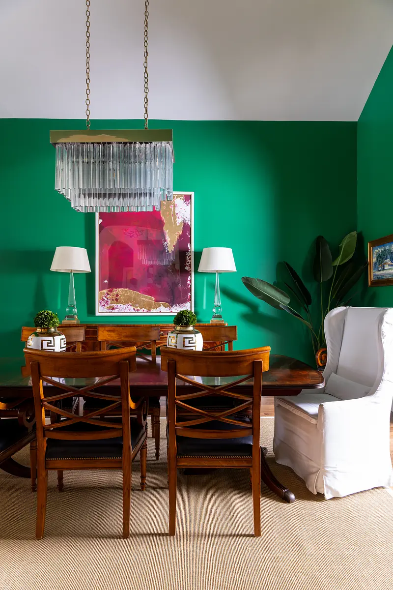 Dining room with wooden table, chairs, white armchair, green walls, artwork, and table lamps.
