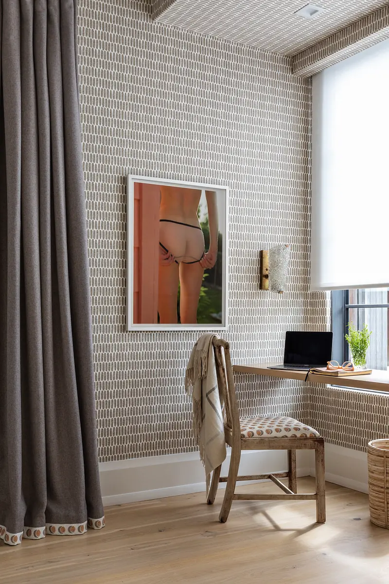 Office area with wooden chair, patterned wall, framed artwork, plant, and curtain beside a window
