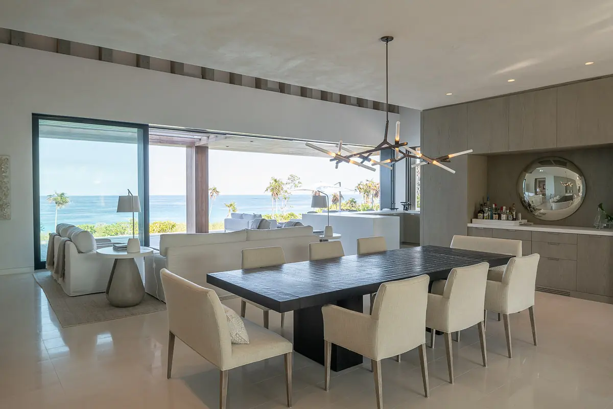 Dining room with a large table, upholstered chairs, ocean view, bar cabinet, and modern chandelier.