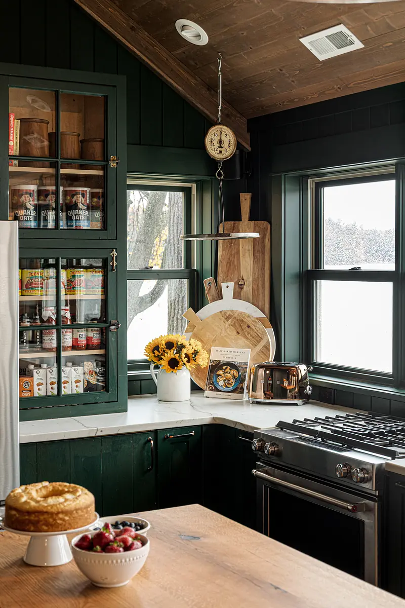 Kitchen with dark green cabinetry, white countertop, gas range, windows, and sunflowers in a vase.
